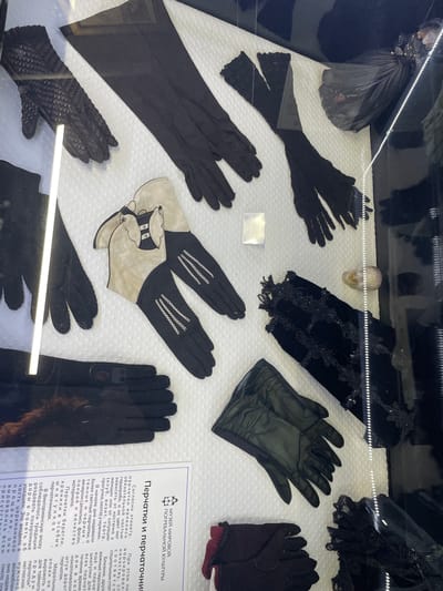 The image displays a museum exhibit featuring a collection of vintage gloves, predominantly in black, arranged on a white textured surface. The gloves vary in material and style, including long, elbow-length gloves, short gloves, and some made of lace or sheer fabric. Some gloves are plain, while others have decorative stitching or embellishments. In the foreground, to the left, a printed informational placard with Russian text is partially visible. The exhibit appears to be housed within a glass display case, as evidenced by reflections on the surface. The overall environment suggests an indoor museum setting. The location of the exhibit is identified as the city of Voskhod, Russia, through the text on the placard. No people are visible in the image, and the objects are static, indicating a curated display rather than an active scene.