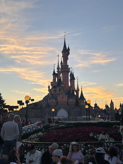 The image shows Sleeping Beauty Castle at Disneyland Paris in Chessy, France, at dusk. The sky is filled with wispy clouds, tinged with orange and yellow from the setting sun, with a contrail visible in the upper portion of the sky. The castle, with its distinctive spires and pink and grey facade, dominates the background. In the foreground, a crowd of people are gathered, some seated and some standing, looking towards the castle or engaged in conversation. Flowerbeds filled with purple and white flowers are visible in the middle ground. Streetlights with glowing orange bulbs are also scattered throughout the scene, illuminating the gathering. The overall atmosphere appears to be one of leisure and observation.