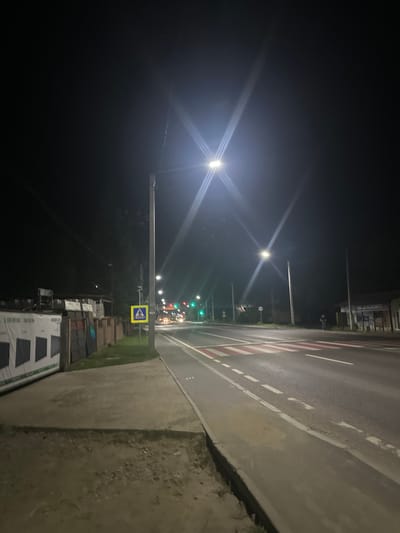 The image captures a street scene at night in the city of Strășeni, Moldova. A pedestrian crossing with red and white markings is visible on the asphalt road. Streetlights illuminate the area, casting bright beams and creating starburst effects due to the camera's aperture. Along the left side of the road, there is a sidewalk, followed by a grassy area and then a fence. Beyond the fence, there are buildings or structures, and some materials that appear to be stacked or stored. Traffic lights are also visible, with a green light illuminated. A pedestrian crossing sign, a yellow diamond with a black silhouette of a person walking, is present near the sidewalk. In the distance, headlights from vehicles suggest ongoing traffic. The sky is dark, indicating nighttime.