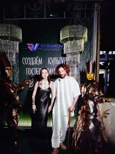 The image shows a red-haired man wearing a white t-shirt and pants, holding a cane, standing next to a woman in a black dress. They are flanked by two figures in elaborate, metallic costumes with golden masks. One costumed figure wears a crown. Behind them is a dark green backdrop with text in Russian, including "RYBAKOV VENTURES" and "СОЗДАЁМ КУЛЬТУРУ" (Creating Culture). Two crystal chandeliers hang from the ceiling. The event appears to be an outdoor or semi-outdoor gathering in a formal or artistic setting, possibly a red carpet event or a promotional display. The location is specified as посёлок дачного хозяйства «Архангельское» (Dacha Settlement "Arkhangelskoye"), Russia.