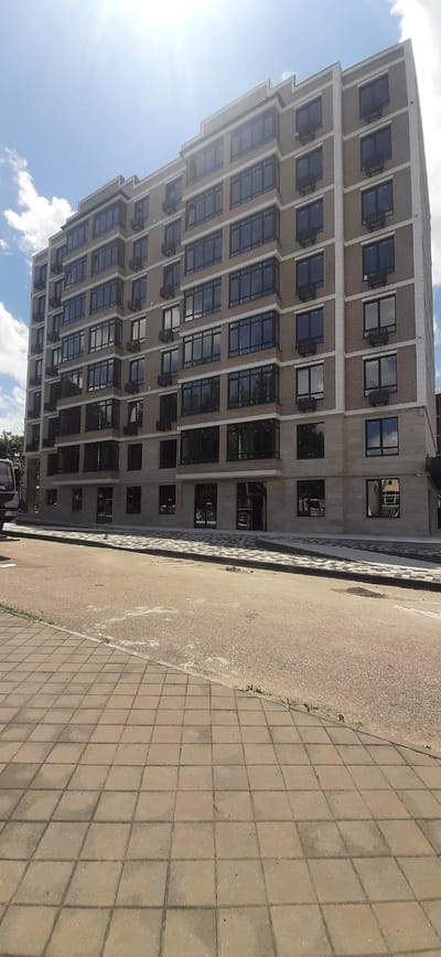 This is a full-frame, eye-level, outdoor, vertical, slightly wide-angle shot of a modern, multi-story apartment building in Yessentukskaya, Russia, on a sunny day with scattered clouds. The building, constructed with light brown brick and light gray stone accents, features numerous dark-framed windows, some with visible air conditioning units mounted below them. The building has a stepped facade on the upper levels. In the foreground, a paved area transitions from light brown cobblestones on the left to a light gray asphalt road surface with white markings. A patterned concrete curb with a black and white diamond design separates the road from the sidewalk. The scene appears to be a street view in front of a residential or mixed-use development, with no people or vehicles clearly visible or the main subjects of the image. The overall impression is one of contemporary urban construction in a suburban or town setting.