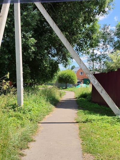 The image shows a paved pathway leading through a grassy area with trees and a building in the background. The pathway appears to be in a rural or suburban setting. On the left side of the pathway, there are two concrete poles supporting a diagonal concrete beam. The area to the left of the pathway is overgrown with tall grass and some smaller plants. To the right of the pathway, there is also grass and a brown corrugated metal fence. In the distance, a two-story brick building with a grey roof is visible, along with some green structures and trees. The sky is blue with some scattered clouds. The overall impression is a sunlit day in a somewhat untended outdoor area.