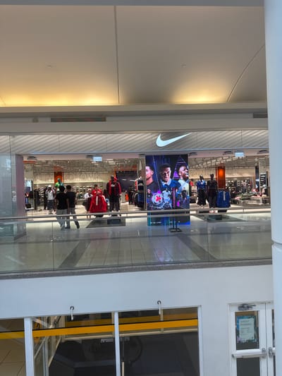 The image depicts the interior of a Nike store, likely located in a shopping mall, possibly in Burnaby, Canada, given the "VANCOUVER" signage visible within the store. The store features a large illuminated Nike logo at the top of its facade. In the foreground, several mannequins are displayed, some wearing athletic apparel. There are also shoppers visible inside the store, browsing merchandise and interacting with each other. A large digital screen displays images of soccer players. The store is well-lit, and the overall environment suggests a commercial setting. The perspective is from an upper level, looking down into the store, with a glass railing in the foreground.