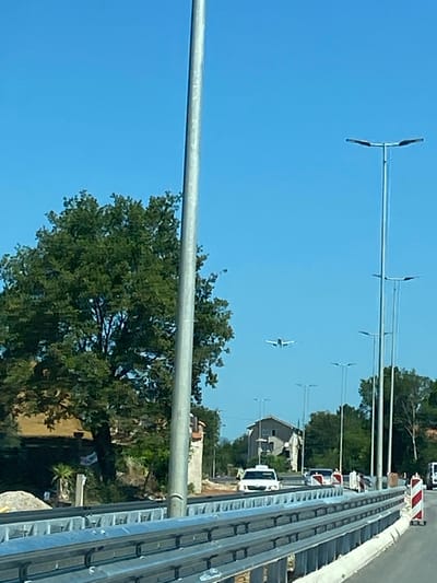 In the city of Radanovići, Montenegro, a clear blue sky dominates the scene. In the distance, a small airplane is visible. A large green tree stands to the left, and further right, a residential building is partially obscured by foliage. In the foreground, a metal guardrail runs horizontally across the frame, indicating a road or bridge. Several streetlights are positioned along the roadside. A white vehicle, possibly a taxi, is driving on the road. Other vehicles and traffic markers are also present. The overall impression is a roadside view on a bright, sunny day.