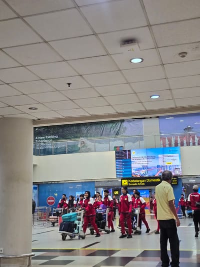 The image depicts the interior of an airport terminal, specifically within the domestic arrivals area. A group of individuals, predominantly young adults, are seen wearing matching red and white athletic uniforms, suggesting they are a sports team or delegation. They are pulling luggage carts laden with bags, indicating they are arriving or departing.
In the background, a large digital display board shows flight information and advertisements. One prominent advertisement displays "Kualanamu Airport: A New Exciting Experience". Another section of the board advertises "Terbang Langsung dan Hemat! Ke Singapura Bersama Scoot!", which translates to "Fly Direct and Save! To Singapore with Scoot!". The overall environment is brightly lit, with a tiled ceiling and floor, characteristic of a modern airport. The atmosphere appears busy, with other travelers and airport personnel visible in the periphery. The presence of airport signage like "Kedatangan Domestik Domestic Arrivals" confirms the location as an airport's arrival hall. The overall scene captures a moment of transit, with the group of uniformed individuals seemingly processing their arrival or preparing for departure.