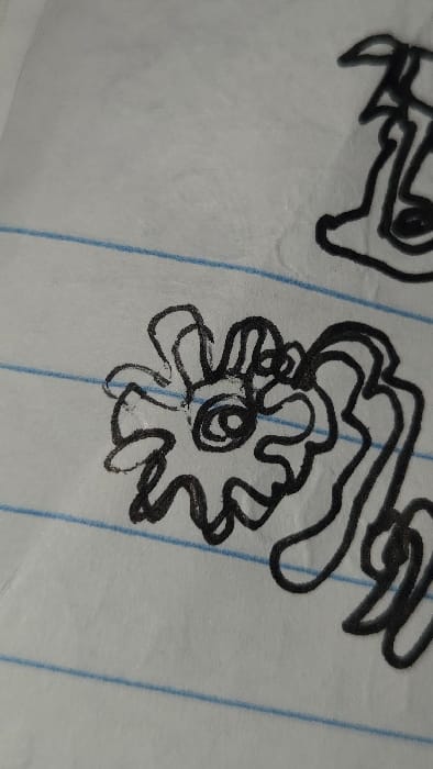 A quick sketch in black ink on lined paper depicts two abstract objects. The first object is partially visible in the upper left, with loosely formed curvy lines. The second object is on the right, and resembles a flower with a circular center, and a cloud-like shape above it. The paper's horizontal blue lines run parallel to the bottom edge.