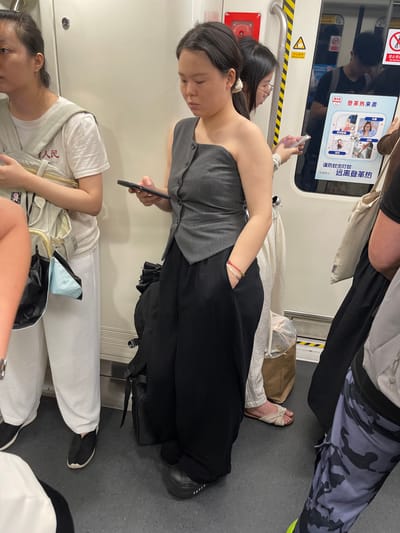The image depicts the interior of a subway car in China. Several people are visible, some sitting and others standing or leaning. One person in the foreground is wearing a grey vest over a black top and black pants, holding a smartphone. Another person, partially visible, is wearing glasses and a white shirt, also looking down at a phone. A third person is seen from the waist down, wearing white pants and black split-toe shoes. In the background, a person in camouflage pants and a black shirt is also present.
Various objects are scattered around, including a brown paper bag, a light blue bag, and luggage. A digital advertisement is displayed on a screen, showing information about preventing dengue fever. Visible text on the advertisement includes "登革热来袭" (Dengue fever is coming), "谨防蚊虫叮咬" (Beware of mosquito bites), and "远离登革热" (Stay away from dengue fever). Other signs in the subway car include "禁止倚" (No leaning) and "小心夹手" (Mind your hand). The overall atmosphere appears to be a typical commute on public transportation. The lighting suggests it could be daytime or artificial lighting within the subway.