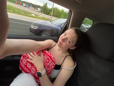 A young woman is lying in the backseat of a car, holding a red and white patterned pillow. She is wearing a black and white tank top and gray pants. She is looking at the camera. The car is parked next to a road with trees and buildings in the background. A white and red pole is also visible. The road has signage in Russian. This scene is located in Staraya Kupavna, Russia.