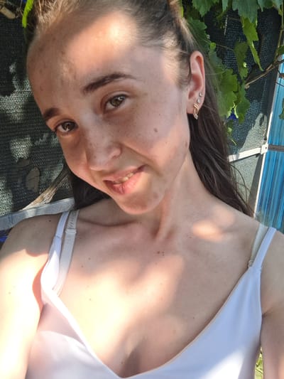 This is a selfie of a young woman lying down outdoors, looking up at the camera. She has brown hair and brown eyes, with freckles across her nose and cheeks. She is wearing a white tank top. Sunlight casts shadows on her face and body. The background is blurred but appears to be a green, leafy vine and a dark, textured surface. There are some blue lines visible at the bottom, possibly indicating a pool or a blue tarp. The overall impression is casual and relaxed, possibly taken on a sunny day in a garden or by a body of water. The woman appears to be smiling faintly, with a relaxed expression. The setting suggests it is daytime, likely late morning or early afternoon given the strong sunlight and shadows.