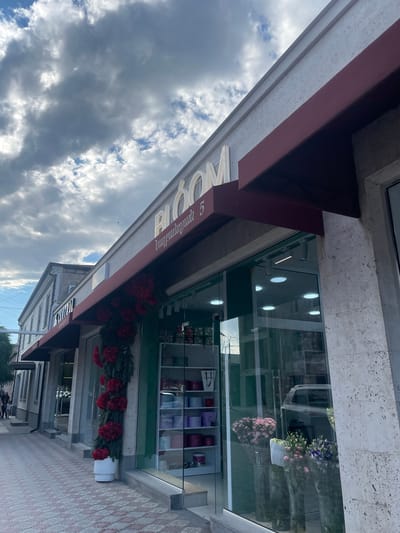 This media file shows a street-level view of a flower shop in Armavir, Armenia. The shop has a large glass storefront displaying bouquets of pink and purple flowers, as well as shelves filled with floral arrangements and gift boxes. Outside the shop, a maroon awning extends over the sidewalk, adorned with red floral decorations and evergreen branches. The building features a stone facade with a textured finish. The sidewalk is paved with a pattern of light and dark pink tiles. The sky above is filled with dramatic clouds, suggesting a dynamic weather condition, possibly late afternoon or early evening given the lighting. The image is oriented vertically, with the sky appearing at the top and the street at the bottom. Visible text on the building includes the name "BLOOM" in large, illuminated letters and the number "5" below it, possibly indicating the street number. There are no people visible in the frame.