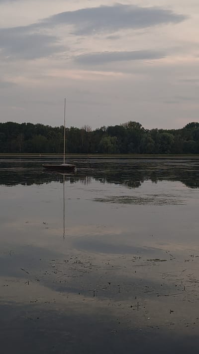 The image depicts a calm lake with a small sailboat anchored in the foreground. The sailboat is dark-hulled with a tall mast. The water reflects the overcast sky, which is filled with soft, gray clouds. A dense line of green trees forms the background, separating the water from the sky. The overall atmosphere is serene and quiet. This scene is located at the Renzenhofer Weiher in Rennertshofen, Germany. There are no people visible in the image.