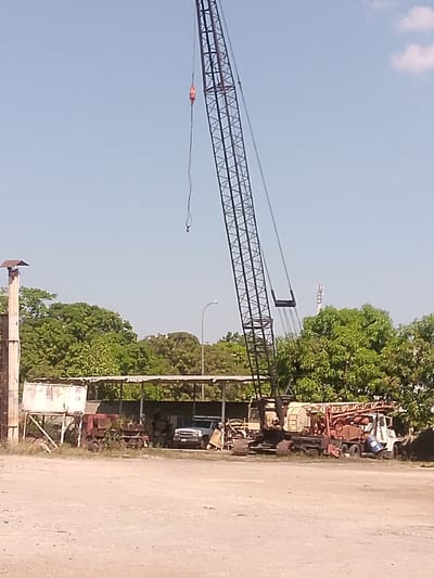 The image shows an industrial scene in Parroquia Guatire, Venezuela. A tall black crane dominates the frame, positioned amidst a collection of parked vehicles and equipment. A weathered open-air structure provides shelter for some items, including a pickup truck. Lush green trees frame the scene, partially obscuring a communications tower in the background. The ground is covered in dirt and gravel, suggesting an active or recently used industrial area. There are no people visible in the image.