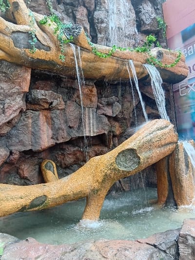 The image shows a man-made waterfall flowing over a large, artificial log in a park in the Pridelands area of Nairobi, Kenya. The log is painted a light brown color and is adorned with vines. The waterfall is cascading from a rock face behind the log, creating a small pool of water at the bottom.  A portion of a building is visible to the right side of the waterfall, but no people are visible in the scene. The water appears to be flowing freely and the rocks surrounding the waterfall appear to be dry. The environment is relatively clean with no visible debris or trash.