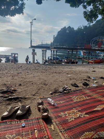 This image captures a scene on the sandy shore of Lake Toba in Parapat, Indonesia, under a bright, partially cloudy sky. The foreground is dominated by a red and black woven mat with floral patterns, on which a pair of beige slip-on shoes with gold buckles are placed. To the right of these shoes, on the same mat, are a pair of brown sandals and a clear plastic water bottle. Further to the right, on the sand, a pair of black flip-flops are visible.
The sand is littered with debris, including dried leaves, plastic bottles, and other small trash items. Beyond the immediate foreground, a sandy beach stretches towards the water. Several people are present in the mid-ground. On the sand near the water, a woman in a dark top and shorts walks away from the camera, while a small child in a yellow shirt walks ahead of her. Further left, near some boats, a group of people are gathered, some sitting and others standing.
The water of Lake Toba is calm and reflects the sunlight, creating a shimmering effect. A wooden pier extends into the lake, supported by pilings. Boats of various sizes and colors are docked or moored near the pier, including a bright orange boat, a boat with a blue and white canopy, and other smaller vessels. Behind the pier, the lush green, tree-covered hills of Parapat rise steeply. The scene appears to be a recreational area, likely a popular spot for locals and tourists to enjoy the lake.