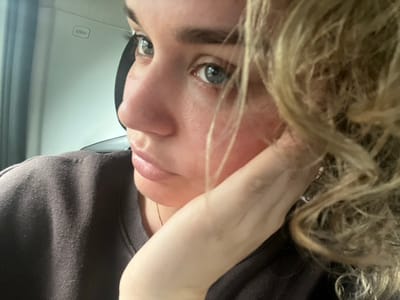 The image shows a close-up of a young woman's face, from the chest up. She is sitting in a car, the back of the driver's seat visible behind her. She has long, blonde hair that is covering part of her face and neck. The woman's eyes are open and she is looking to the left side of the image. Her mouth is slightly open and her lips are slightly parted. She is wearing a dark brown, knitted sweater. The woman's right hand is resting on her left cheek, and her left hand is on her lap. The image appears to have been taken indoors in a car. The location of the image is Blåvand, Denmark, based on the provided coordinates.