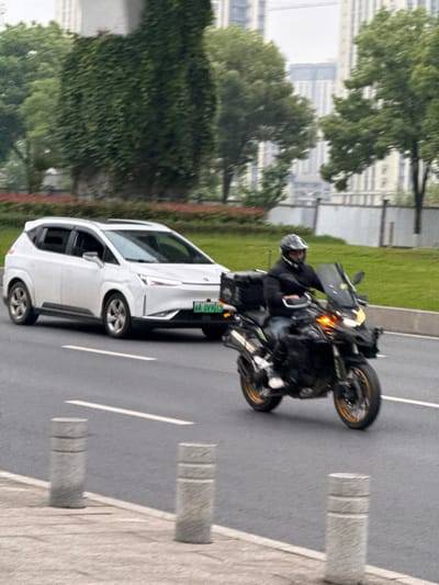 This image captures a dynamic street scene in Shuangfu, China, under what appears to be an overcast sky.

In the foreground, a dark-colored motorcycle is prominently featured, moving along a multi-lane asphalt road. The motorcycle has distinctive gold-colored wheel rims and its front amber lights are illuminated, possibly functioning as turn signals or daytime running lights. The rider, wearing a helmet and a dark jacket, is visible and appears to be in control, with a slight motion blur around them and the vehicle, indicating movement.

Adjacent to the motorcycle, a white SUV is also in motion. A notable detail on the car is its green license plate, which reads "GA DV901S." This green plate typically designates a new energy vehicle (such as an electric or plug-in hybrid car) in China.

The road features clear white lane markings. To the right of the road, there's a light-colored paved area, likely a sidewalk, lined with several grey cylindrical bollards. Beyond the immediate roadside, the environment includes lush green grass and trees, suggesting a well-maintained urban or suburban landscape. In the background, partially obscured by the foliage, are several multi-story buildings, contributing to the urban setting.

The diffused lighting indicates an overcast day, with no strong shadows evident. The overall impression is one of vehicles traveling through a contemporary Chinese city.
