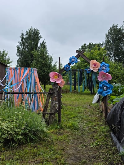 The image depicts an outdoor scene with a rustic wooden archway decorated with large, colorful paper flowers in shades of pink, peach, and blue. Strands of orange, white, and blue ribbons hang from a frame to the left of the archway, creating a decorative curtain. The ground is covered in green grass and appears damp. Lush green trees and foliage surround the area. In the background, a wooden structure resembling a shed or fence can be seen. The sky is overcast, suggesting a cloudy day. The scene appears to be set in a rural or garden environment, possibly in the village of Старые Быги, Russia. There are no people visible in the image.