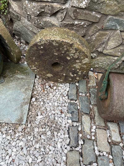 The image shows a ground-level view of a stone-paved area in Minsterley, United Kingdom. A roughly hewn stone wall borders the left side of the frame. Adjacent to the wall is a large, circular millstone lying on its side, featuring a prominent central hole. To the right of the millstone, the ground is covered with small, loose gravel. A section of paving stones is visible in the upper right portion of the image. A rusty metal object, possibly another millstone, is partially visible in the top right corner, attached to a green metal arm or handle. Pine needles and other small debris are scattered throughout the gravel and paving stones.