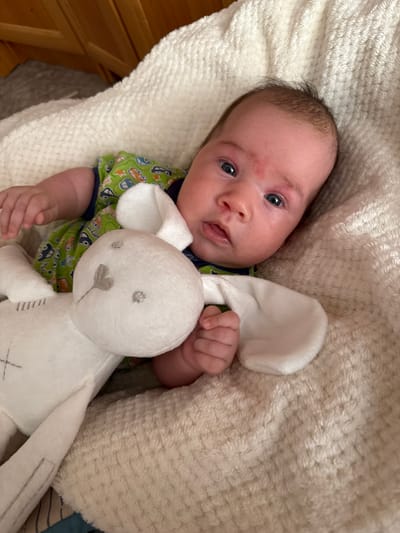 A close-up overhead shot shows a newborn baby lying on a fuzzy, cream-colored blanket. The baby is facing upwards with their head slightly turned to the left. They have fair skin, sparse dark hair, and wide blue eyes. There's a red mark on their forehead, possibly a birthmark or rash, and another smaller red mark near their left eye. The baby is wearing a green short-sleeved onesie with a pattern of cartoon cars and stars.
In the baby's right hand, they are holding a white plush bunny toy. The toy has embroidered grey features on its face, including eyes, a nose, and a mouth, and a stitched heart shape on its chest. The baby's left hand is also visible, resting on the blanket.
The background is dominated by the soft texture of the blanket. To the left, a wooden surface with a diagonal grain is visible, suggesting furniture or a wall. The lighting appears to be indoor, with no indication of time of day or weather. The overall scene conveys a sense of peace and comfort. No text is visible in the image. The location context is Выльгорт, Russia, but this information cannot be visually confirmed from the image alone.