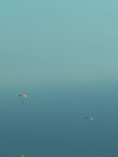 The image depicts a clear blue sky with two paragliders actively engaged in their sport. The paragliders are visible as small figures against the vast expanse of the sky. One paraglider, featuring a blue and yellow wing, is positioned higher in the frame. Below and to the right, another paraglider with an orange wing is visible, accompanied by a person.
The scene is set outdoors, with the sky appearing calm and clear, suggesting fair weather conditions. The lighting indicates it could be daytime, possibly late afternoon or early evening given the soft quality of light on the paragliders' wings. There are no discernible location cues within the image itself, but the context provided places the activity in Ivanovići, Montenegro. The overall impression is one of freedom and exhilaration associated with paragliding. No text is visible in the image.
