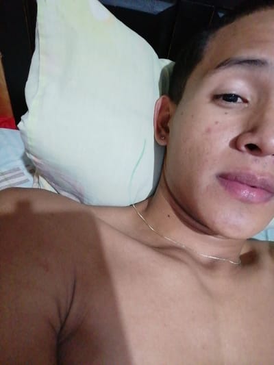 A close-up shot shows a person lying on a bed with a white pillow. The individual has short dark hair, brown eyes, and a fair complexion with some skin blemishes. A thin gold chain necklace is visible around their neck. The person's upper body is bare, exposing their shoulders and chest. The bed has blue and white patterned sheets. The background includes a dark headboard.
