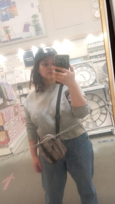 The image depicts a woman taking a selfie in a store setting. She is wearing a gray sweatshirt, blue jeans, and a baseball cap. A small gray bag hangs across her body. The background reveals store shelves stocked with various items, including clocks and home decor products. Based on additional information, the location is Юдино, Russia.