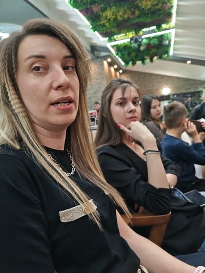 The image depicts an indoor scene within a public establishment in Opaka, Bulgaria. In the foreground, a female subject with light brown hair featuring distinctive crimped sections, appearing to be in her 30s-40s, is shown from the chest up, facing slightly right with her mouth partially open. She wears a black long-sleeved top, a silver-toned chain necklace, and a rectangular, woven gold-toned brooch on her left chest area.

Behind her, to the right, a younger female subject, likely in her 20s, with dark hair, is seated and looks directly at the viewer, resting her chin on her left hand. Her fingernails are painted red, and she wears a black top and a dark bracelet on her left wrist. Further back, another female subject with dark hair is partially visible, followed by a young male in a blue shirt with his hand near his ear. Distant figures are faintly present in the background.

The environment includes textured grey and brown walls. Overhead, large rectangular structures adorned with artificial green foliage and colorful accents are suspended from a white ceiling, incorporating linear lighting elements. Seating visible is constructed of wood.