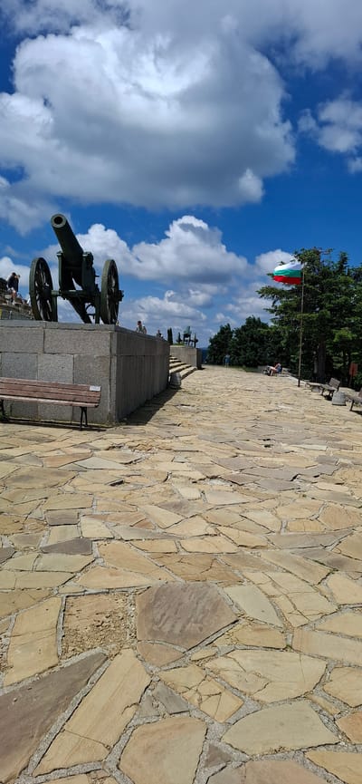 The image depicts a view of the Shipka Memorial, located on St. Nicholas Peak near the town of Shipka, Bulgaria. The memorial complex features a large, old cannon mounted on a stone pedestal in the foreground. The cannon is green and appears to be made of metal, with large spoked wheels. Behind the cannon, there are tiered stone steps and pathways paved with irregular flagstones. Several people are visible in the distance, some walking and others seated on benches. A Bulgarian flag, with white, green, and red horizontal stripes, is flying from a flagpole to the right of the memorial. The sky is blue with scattered white clouds. The environment is outdoors, with lush green trees visible behind the flag. The overall scene suggests a historical monument and a place of historical significance.