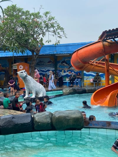 The image shows a waterpark in Kecamatan Tanggulangin, Indonesia, located at coordinates -7.497 latitude, 112.684 longitude. The park features a large swimming pool with a bright blue tiled surface. There is a tall orange waterslide to the right of the pool, which is constructed of metal and plastic, leading down into the pool. There are several people in the water, as well as a few seated on rocks surrounding the pool. There are some people standing around the edge of the pool as well. The pool is partially enclosed by a low wall and surrounded by large rocks. The park is decorated with painted murals on the surrounding building walls featuring sea creatures and underwater scenes. Several trees and lush vegetation provide shade over the park.  A white polar bear statue stands on a platform between the pool and the wall, facing the pool. The image was taken during the day. There is a clear sky in the background. The image is slightly blurred, but it is clear enough to identify the location.