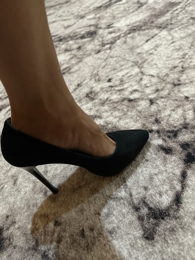 A close-up shot focuses on a woman's left foot, adorned in a black, pointed-toe stiletto heel, positioned on a textured carpet with a grey and white marble-like pattern. The shoe's heel is thin and metallic, casting a distinct shadow on the carpet. The carpet exhibits a soft, plush texture. The image does not provide any visual cues to determine the location within the city of Kalininskaya, Russia, or any specific context beyond the immediate subject and its surroundings.