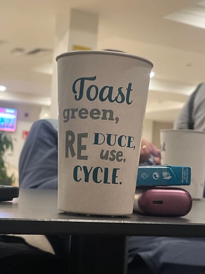 The scene is set indoors, likely within a lounge, cafe, or hotel lobby in Calvià, Spain. A beige reusable cup, printed with the text "Toast green, REDUCE, use, CYCLE" in blue and grey lettering, dominates the foreground, positioned on a dark table surface. To the right of this cup, a small, light blue rectangular packet, consistent with a pack of cigarettes or gum, displays recycling symbols and unreadable foreign text. Immediately beneath this packet, a maroon, oval-shaped wireless earbud charging case, featuring a visible charging port, rests on the table. In the blurred midground, a person's legs in blue trousers are visible to the left. To the right, a second white cup is partially visible, alongside a hand wearing a ring and a blurred figure in a light-colored garment. The background reveals a light-colored ceiling with embedded light fixtures, a television screen displaying colorful content on the left, and faint green foliage. The area is illuminated by overhead artificial lighting.