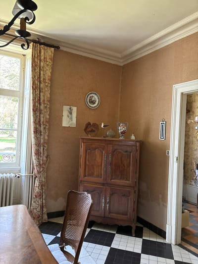 The image captures an indoor scene, likely a room within a traditional home in Étréchy, France, during daylight hours. The room features textured, light brown wallpaper, complemented by white crown molding and a white ceiling.

Dominating the right side of the room is a large, antique dark wood cabinet or armoire, adorned with ornate metal handles and carved panels. In front of it, a chair with a cane or wicker back and a wooden frame is partially visible, along with a wooden table in the foreground.

Decorative items on the wall include a circular plate with a floral pattern and a small framed picture (possibly a sketch) beneath it. On top of the cabinet, several smaller decorative objects are arranged, including a glass vase or bowl, a small white bird figurine, and a dried botanical piece.

A large window, framed in white, occupies the lower part of the left wall, looking out onto a lush green outdoor area. A patterned curtain, featuring a floral design with red and pink flowers on a beige background, hangs over the window. A white radiator is visible beneath the window.

In the upper right, an open doorway reveals an adjoining space with a striking black and white checkered tiled floor and patterned wallpaper. A rectangular vent is visible on the wall near the ceiling. A dark metal wall sconce or light fixture is partially seen in the bottom left corner.

The room is empty of people, suggesting a quiet, unoccupied space. No discernible text is visible within the image.