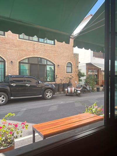 An outdoor urban scene captured from an elevated perspective shows a brick building with arched windows and an upper-level round window. A black pickup truck is parked along the road adjacent to the building. A bench with a wooden seat stands nearby, framed by pink flowers in a pot. Two parked scooters are visible further down the road. A green awning stretches over the top left of the frame, and a partially visible window frame outlines the right edge of the image.