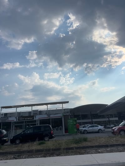 This media file shows a scene with a cloudy sky and a building with cars parked in front of it. The image is oriented vertically, with the sky occupying the top portion and the ground with the building and cars below. The sky is partly cloudy, with patches of blue sky visible and sunlight filtering through the clouds, suggesting it is daytime. The weather appears to be mixed, with both sun and clouds present.

The building appears to be a commercial establishment, possibly a restaurant or a business with multiple units, given the signage visible. One visible sign says "CORNER RESTAURANT." There are several cars parked, including a black van, a white car, and a red car. One person, wearing a red shirt, is visible near a green ATM machine.

The scene is set in a location that appears to be an urban or suburban area with a road and a building. The grass and concrete surroundings suggest an outdoor setting. There are no specific clues about the exact time of day beyond it being daytime, and no strong emotional cues are conveyed by the image. The overall impression is a snapshot of a day in what seems like a commercial or service area. The location context provided is Dub, Montenegro.