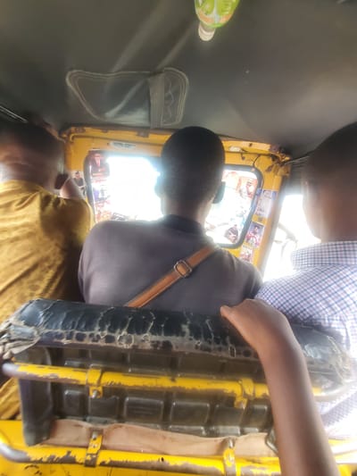 The image shows the interior of a yellow auto-rickshaw, likely a "Keke" or "Maruwa," typical of Nigeria, specifically in the city of Ojoto. Several passengers are visible. One wears a light blue and white checkered shirt, sitting next to a person in a grey shirt with a brown strap across their shoulder. Another passenger wears a yellow shirt. The auto-rickshaw's interior is decorated with stickers on the window. The vehicle appears to be in motion, with a bright light source visible outside the window. The seat in the foreground is worn and damaged.