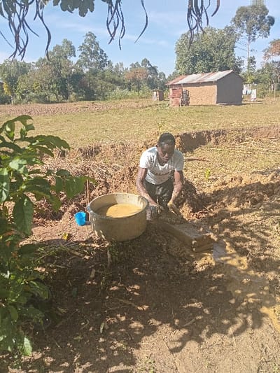 A man in a white t-shirt crouches near a ditch, next to a tan bucket. The surrounding landscape includes a grassy field, trees, and a small building with a metal roof. Patches of sunlight and shadows cover the ground. Some green bushes are near the camera. The sky is blue.