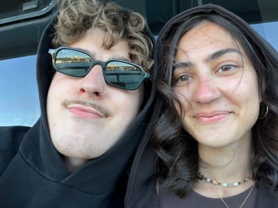 Two people, a woman and a man, are shown in a vertically oriented, close-up shot, apparently lying down. The woman is on the left wearing a brown hooded top, while the man is on the right in a black hoodie and sunglasses. The woman has long dark hair and a beaded necklace with a gold chain. The man is wearing a nose ring, has light brown hair, and his sunglasses reflect a waterfront view. The reflection shows buildings along the shoreline in what appears to be Kadıköy, Turkey. The background is a slightly blurred blue-grey surface.