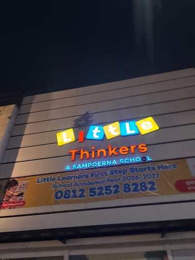 The media file shows a brightly lit sign for "Little Thinkers A SAMPOERNA SCHOOL" at night. The sign features colorful, illuminated letters spelling out "Little Thinkers" in a playful font, with "A SAMPOERNA SCHOOL" written below in a more standard font. A yellow banner is attached to the sign, displaying text that reads "Little Learners First Step Starts Here," "School Academic Year 2026-2027," and a phone number: "0812 5252 8282." The banner also has small images of children's books or learning materials, and a partial image of a child. The overall impression is that of an educational institution aiming to attract young learners. The setting appears to be an outdoor area in front of a building, likely in South Tangerang, Indonesia, given the context provided. The time of day is clearly night, indicated by the dark background and the illuminated sign. There are no visible weather conditions mentioned or depicted. The image focuses on the signage itself, with no prominent depiction of people or specific activities beyond what is represented on the banner.