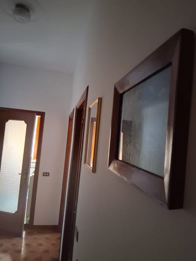 The image displays an interior scene, focusing on the ceiling and a doorway. Dominating the upper portion of the frame is a large, square skylight with a dark wooden frame, offering a distorted reflection of what appears to be the outdoors or an upper level, possibly depicting a cloudy sky or a textured surface. Below the skylight, a rectangular mirror with a golden frame is suspended, also reflecting light. Further down, a more elaborate structure, possibly part of a decorative ceiling feature or another framed opening, with a dark wooden frame and an orange interior is visible.
In the lower half of the image, a wooden door with a textured glass panel is partially open, allowing light to stream through. The door appears to have a metal handle. Next to the doorway, a light switch with two outlets is mounted on a light-colored wall. To the left of the doorway, a circular light fixture with a textured cover is attached to the wall. The overall lighting suggests an indoor environment, potentially during daylight hours given the light entering through the door. The scene seems to be a residential or interior space, with elements of architectural detail and decoration. There is no visible text in the image.