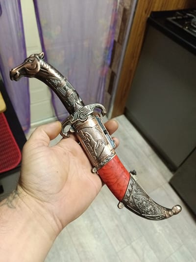 A close-up shot shows a person's hand holding a decorative dagger. The dagger has an ornate, bronze-colored handle shaped like a horse's head with intricate patterns along its neck. The hilt features detailed metalwork with a depiction of a horse and rider. The handle is wrapped in a textured, reddish material, possibly coral or faux coral, and capped with a decorative metal pommel. The blade is partially visible and appears to be metallic. The background is blurred, showing hints of purple curtains, wooden surfaces, and a dark countertop. The image is taken in the city of Yukariyuz, Uzbekistan.