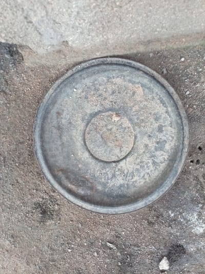 An overhead, medium shot shows a rusty, circular metal object, possibly a pot lid or manhole cover, resting on a patch of gritty, sandy soil. The object is dark gray and shows signs of wear and tear, with concentric circles etched into its surface. The soil around the object is a mix of light and dark brown sand, with small pebbles and some dark debris scattered throughout. The background is a rough, textured surface that appears to be concrete or stone, with a lighter gray hue. The scene appears to be outdoors in a dry, possibly dusty environment. There are no people or animals visible in the image. The location is not specified in the image.