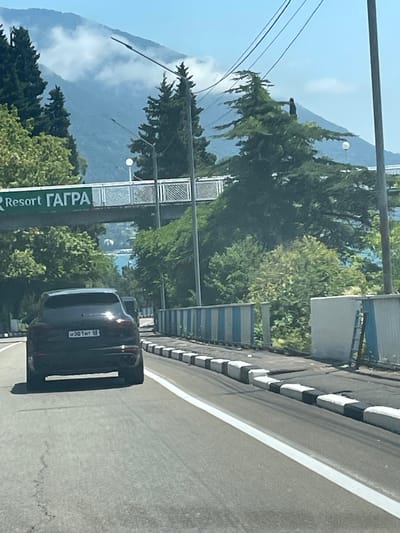 A black SUV drives on a paved road in Abaaata, Abkhazia, beneath a clear blue sky and distant, cloud-shrouded mountains. The SUV is in the foreground, facing away from the camera, with its license plate visible. A green sign with white lettering reading "Resort ГАГРА" is suspended over the road in the middle ground. Trees line the sides of the road, and a body of water is visible in the distance beyond the trees. A pedestrian overpass with a metal railing crosses the road in the upper middle ground. A striped barrier, alternating between blue and white, runs along the right side of the road.