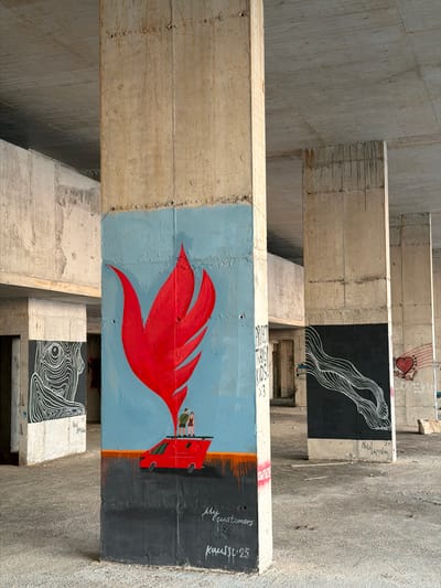 The image depicts an urban environment under a concrete overpass, likely in Perazića Do, Montenegro, characterized by its unfinished concrete structure and weathered surfaces. The focal point is a large concrete pillar adorned with colorful graffiti art.
On the main pillar, a vibrant red, flame-like motif dominates the upper portion, set against a bright blue background. Below this, a red van is depicted with two small figures standing on its roof. The lower section of this pillar is painted black, with white cursive script that appears to read "my customers" and "Kassu '25".
To the left, another pillar features a black and white mural of a stylized human profile composed of concentric white lines. To the right, a pillar has a black background with a white wavy line pattern. Further to the right, another pillar displays a red heart with stylized flag-like stripes behind it, and text that appears to read "LOVEKU". The ground is concrete, with scattered debris. The overall atmosphere is one of urban decay and artistic intervention.