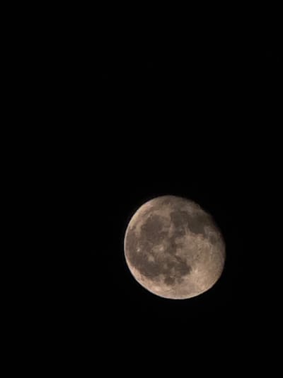 A full moon is visible in a dark sky, occupying the lower right portion of the frame. The moon's surface shows craters and maria, with variations in light and shadow creating a textured appearance. The background is entirely black, suggesting a nighttime scene with no other celestial bodies or atmospheric elements visible. The image is a close-up of the moon against a void.