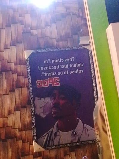 The scene is indoors, located in Enugwu-Ukwu, Nigeria. A vertically oriented rectangular poster, primarily purple in color, is mounted on a wall. The poster displays a mirrored illustration of rapper Tupac Shakur, depicted in profile, facing left, with a plume of smoke emanating from his mouth. He wears a dark cap and a light-colored collared shirt over a white undershirt. Above the illustration, a mirrored quote reads, "They claim I'm violent just because I refuse to be silent." Below the quote, the mirrored name "2PAC" is prominently displayed in red, uppercase letters. The poster features a decorative border around its perimeter and is secured at the corners by small, light-colored triangular fasteners. The wall behind the poster is covered with a textured paneling resembling stacked, irregular wooden planks in varying shades of brown. To the right of the poster, a vertical section of a bright green surface is visible, likely a wall or partition. Adjacent to this green surface on the right, a vertical white structural element is present. A significant glare from an overhead light source is visible in the upper center of the image, reflecting off the poster and the wooden wall.