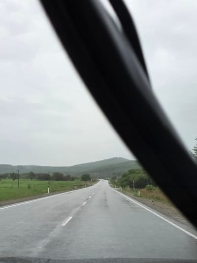 A view from inside a vehicle captures a long, wet asphalt road stretching into the distance under an overcast sky. A dark, blurred object, likely a car dashboard or a pillar, diagonally crosses the foreground from the upper left to the lower right, partially framing the scene. The road, marked with white dashed lines, appears recently rained upon, reflecting the muted light of the sky. Green fields and low-lying vegetation line both sides of the road, with some small posts or markers visible alongside. In the mid-ground, a range of low hills or mountains extends across the horizon, blending into the uniformly gray, cloudy sky. There are no people visible, and the road appears empty. The overall impression is one of a journey through a rural or semi-rural landscape during an overcast and wet day. The location context suggests this scene is in Бабстово, Russia. No discernible text is present in the image.