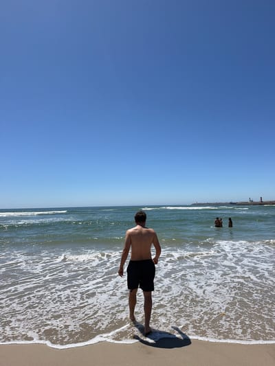 In the city of Gafanha da Nazaré, Portugal, a man stands on a sandy beach with his back to the camera, facing the ocean. He is wearing black swim trunks. The water is a deep green and blue, with gentle waves breaking on the shore, creating a white foamy edge. In the distance, several people are visible in the water, and a breakwater structure extends into the sea, topped with lighthouses. The sky is a clear, vibrant blue, indicating a sunny day.