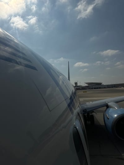The image shows the side of an airplane, taken from within the plane. The airplane is parked at Ben Gurion Airport in Tel Aviv, Israel, which is located at the coordinates 32.002, 34.87. The side of the plane is white with a blue stripe pattern. The blue stripe pattern is a logo, but it is difficult to determine what it is. The wing of the plane can be seen extending out to the right of the frame. Another plane is visible in the distance, partially obscured by a building, as well as a tall control tower. The airport tarmac is visible in the distance, and a blue sky with white clouds can be seen above.