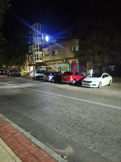 The image depicts a street scene at night in Harmanli, Bulgaria. Several cars are parked along the side of the road, including a white sedan, a red hatchback, a dark-colored sedan, and a silver station wagon. A multi-story building with a distinctive glass-enclosed upper section is visible in the background, illuminated by interior lights. Trees line the street, and the asphalt road is marked with white lines. A brick and concrete sidewalk runs along the edge of the road. The overall impression is of a quiet urban setting at night.