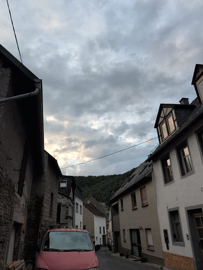 This is a photograph taken in Monreal, Germany, depicting a street scene with buildings and a cloudy sky. The perspective is from a low angle, looking upwards towards the sky and the upper stories of buildings.

The scene shows a narrow street lined with traditional German houses. These buildings have stone and stucco facades, with various window styles, some with shutters. One house features a prominent dormer window with skylights. A pink car is parked on the right side of the street, partially visible, with its windshield reflecting the sky. In the background, a lush green hillside is visible, suggesting a hilly or mountainous terrain surrounding the town. The foreground shows the dark, rough texture of a stone wall and a section of a roof with a gutter.

The sky is filled with dramatic, heavy clouds, with patches of bright sunlight breaking through in some areas, indicating a dynamic weather condition, possibly late afternoon or early evening. The overall atmosphere is somewhat somber due to the heavy cloud cover, but the hints of sunlight add a touch of hope or drama. There is no visible text in the image. The scene captures a slice of daily life in a quaint German village.